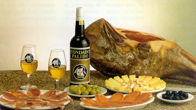 Una botella y dos copas de vino  del Condado  posan en una mesa junto a un jamón  y platos de queso, lomo ibérico y aceitunas.
