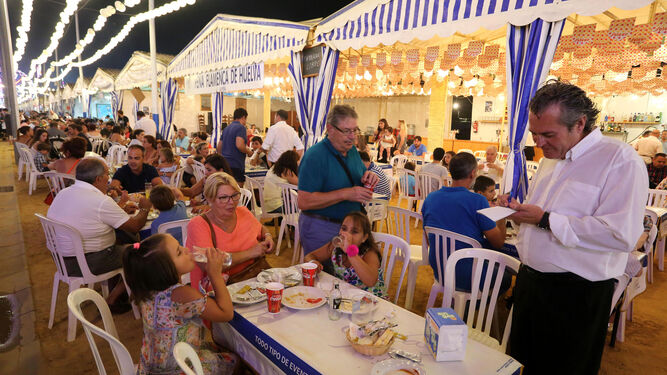 Un camarero atiende a una familia en una mesa en la caseta de la Peña Flamenca de Huelva durante la noche del viernes en el recinto.