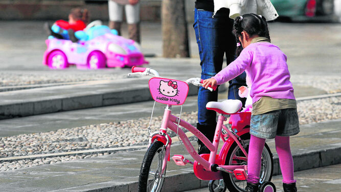 Una niña  juega con su bicicleta en un parque, vigilada por su madre.